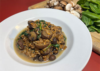 Mushroom and spinach farro risotto is displayed in a white bowl on a red counter with raw spinach and mushrooms on a cutting board.