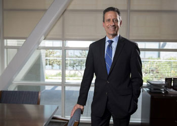 uc irvine school of medicine dean michael stamos wearing a dark suit blue tie standing in his office next to a desk