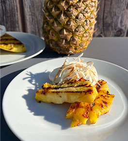 Hawaiian grilled pineapple with whipped coconut yogurt displayed on white plate with whole pineapple and gray fence in background.