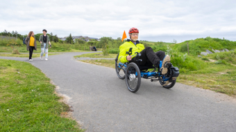 A man with a white beard wearing high-vis using an adapted trike in Arcadia Park in Scotland on a cloudy day