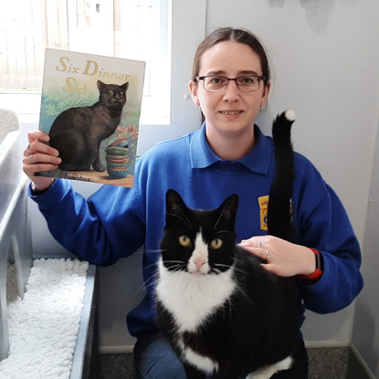 Woman wearing Cats Protection shirt holding Six Dinners Sid book with black cat on her lap