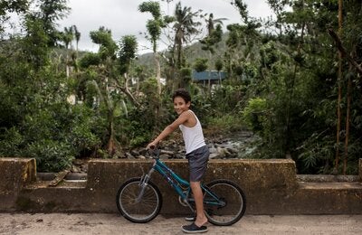 A boy of around 10-years-old is seated on a bicycle with his feet on the ground and his hands holding the bike, over a cement bridge. In the background, a forest of tropical trees and bushes hide the roofs of a building ant the shilouette of a mountain