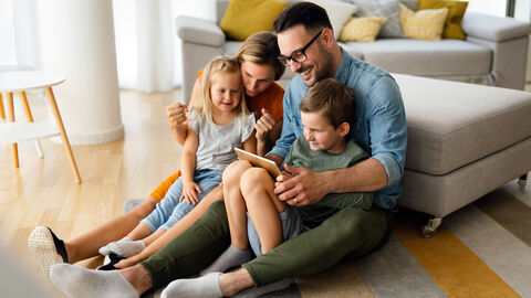 Ein Mann und eine Frau sitzen mit einem Mädchen und einem jungen auf dem Schoß auf dem Boden vor einer Couch und schauen in eine Tablet.