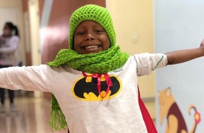 Niño afrodescendiente con los brazos extendidos y una gran sonrisa en su rostro. Vestido con una camiseta con el logo de Batman y un gorro y bufanda tejidos con lana verde