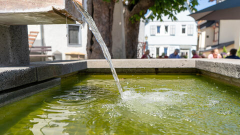 In einem Brunnen fließt Wasser, im Hintergrund sitzen Menschen in einer Gaststätte