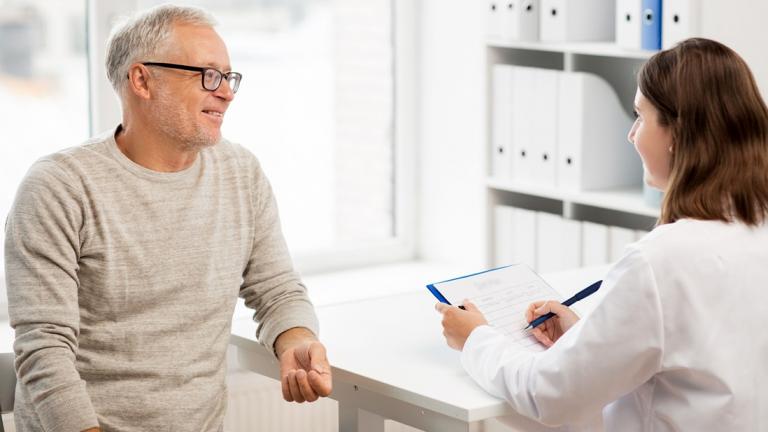 A male patient wearing glasses sits in chair while he speaks to a female healthcare provider about his health.