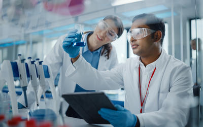 A male researcher in a high-tech medical lab holds up a vial of blue liquid while a female researcher next to him leans in to look at it.