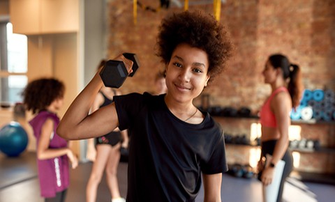 African american boy smiling at camera while exercising using dumbbell in gym together with female trainer and other kids.