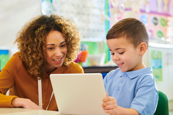 A teacher looks encouraging and engaged with student who is working on a tablet a classroom