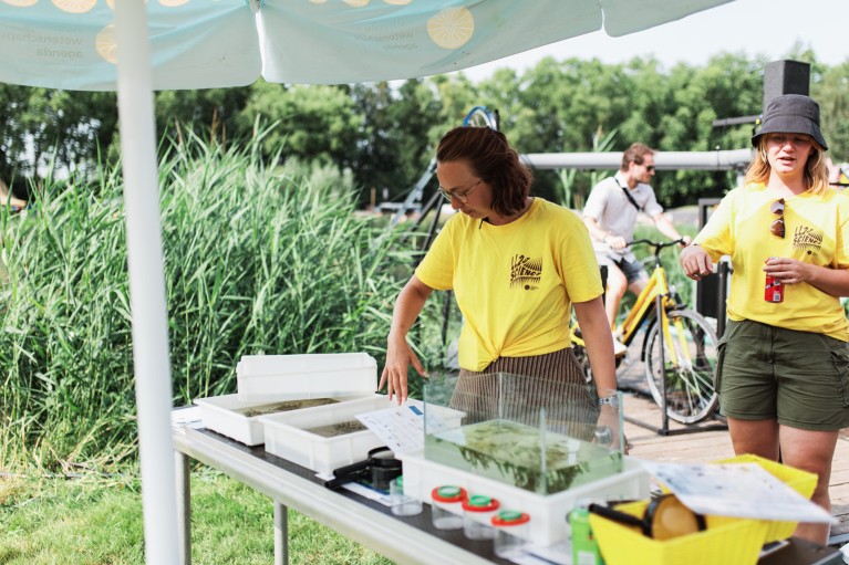 Two people wearing yellow t-shirts stand at a table with square containers of lake water