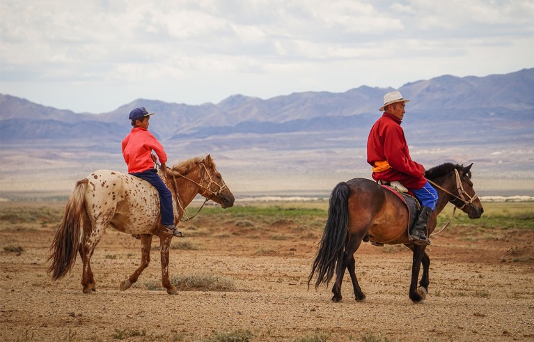 Two men in red shirts sit atop horses; towering mountains stretch across the background.