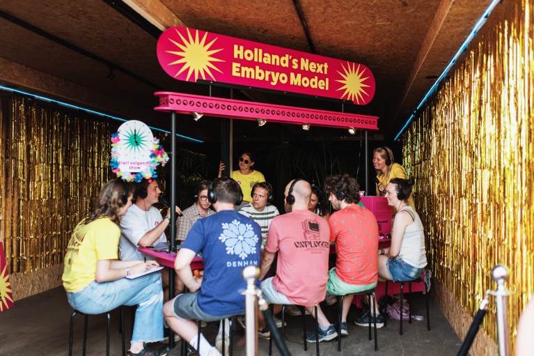 Festival visitors wear headphones and sit at a table with a sign reading 'Holland's next embryo model'