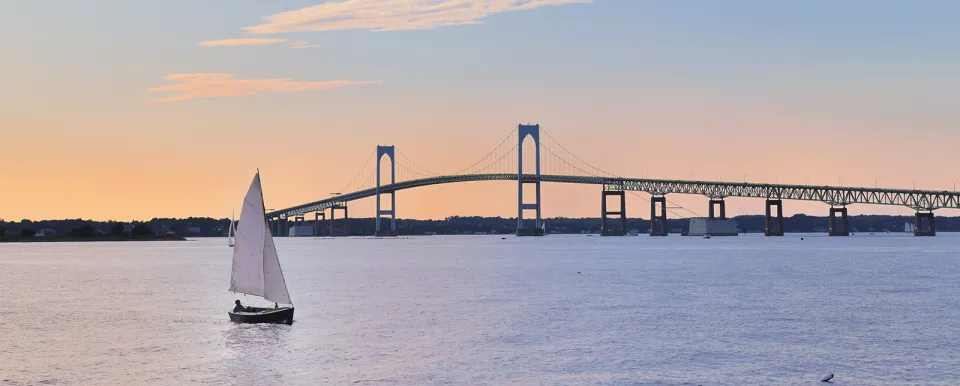 A solo sailor in a small sailboat out on the water under a Rhode Island bridge