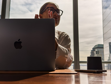 En kvinde sidder ved et bord i et højhus. Bag hende er der et stort vindue med udsigt til andre bygninger. Foran hende er der en åben MacBook.