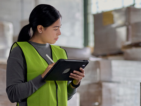 En kvinde står på en fabrik med en iPad i hånden og kontrollerer lagerbeholdningen. Bag hende er der stakkevis af store kasser.