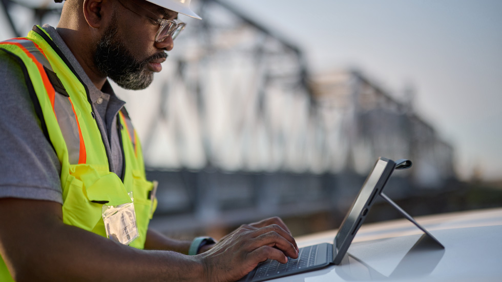 Un homme portant un casque de chantier tape sur le clavier d’un iPad. Il est en extérieur. On aperçoit un pont derrière lui.