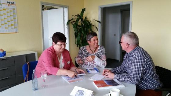 Zwei Frauen und ein Mann sitzen in einem Büro an einem Tisch. Sie haben Unterlagen und Broschüren vor sich liegen und sprechen miteinander.