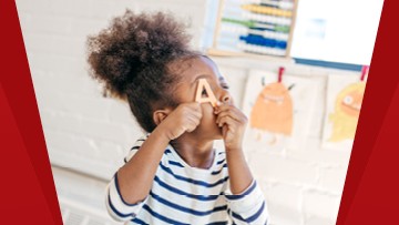 A child is holding the letter “A,” which is made of wood. There are children’s drawings in the background.