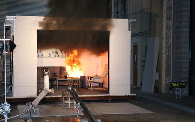 A couch is in flames in a mocked-up room inside a large fire testing facility. 