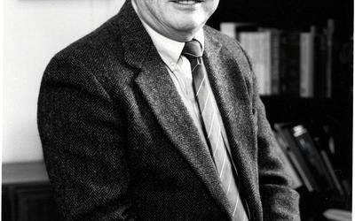 Vertical, black and white image of a man in a suit sitting at the edge of a desk.