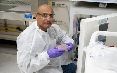 A researcher wearing safety gear kneels at the open door of a lab fridge, displaying a tray of samples. 