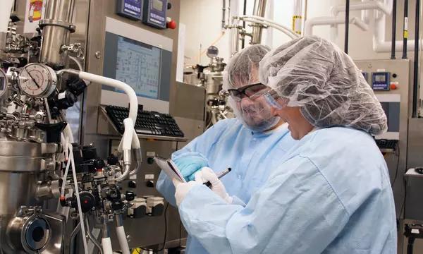 Two people in coveralls and hair covers check the readouts on laboratory equipment.