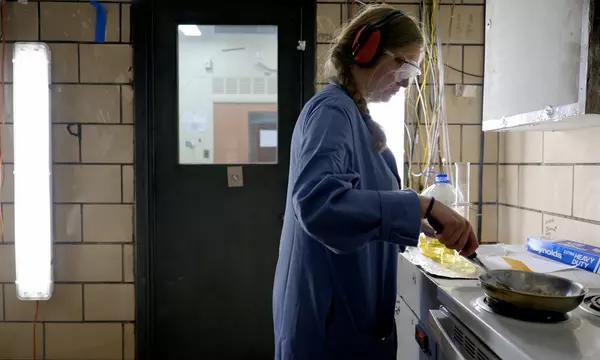 Side view of a woman wearing safety glasses, ear protection, and a blue lab coat. With one hand, she is holding a stainless steel frying pan over an electric stove. With her other hand, she is using a spatula to prod bacon in the pan. The room is smoky.