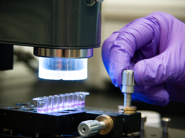 A hand in a purple glove turns a metal dial on a plate holding tiny vials of purple liquid. 