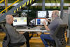 Two men sit at a table in a lab, facing each other and smiling as they talk and gesture toward their computer screens. 