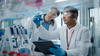 A male researcher in a high-tech medical lab holds up a vial of blue liquid while a female researcher next to him leans in to look at it.