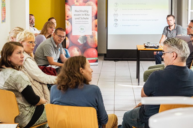 Menschen sitzen in einem Halbkreis vor einer Präsentation mittels Beamer. 