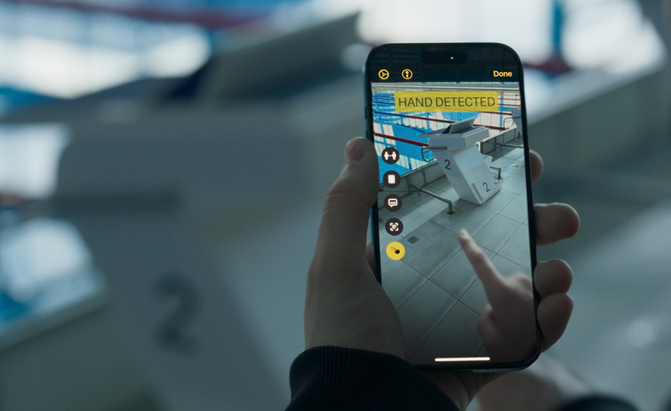 Une main tenant un iPhone, avec l’appareil photo pointé vers un plot de natation. Un doigt est posé sur l’écran de l’iPhone et les mots « MAIN DÉTECTÉE » apparaissent.