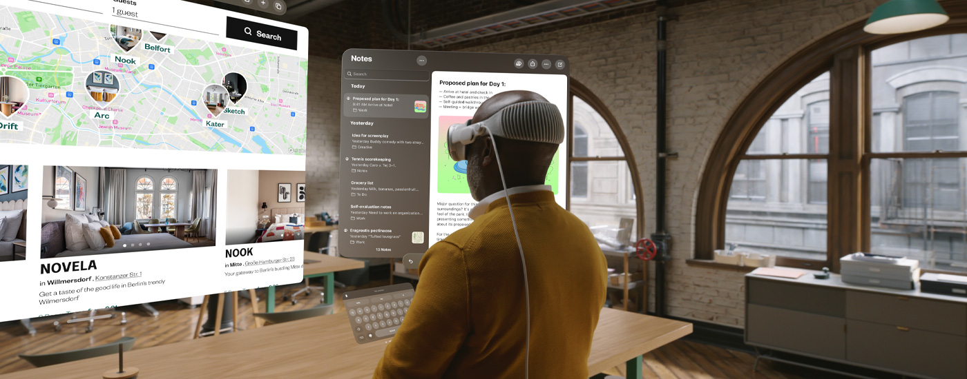 Une vue légèrement de profil d’un homme portant l’Apple Vision Pro, qui regarde plusieurs écrans affichant une carte et l’app Notes. Il se tient debout à son poste dans un espace de bureaux en ville.