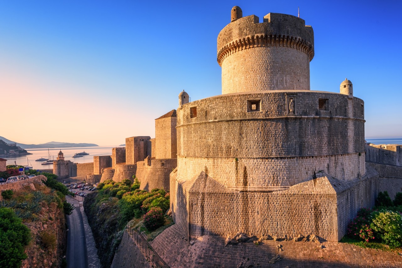 View of a huge, circular fortress with a round castle tower in the centre in the evening light. The fortress is part of a larger fortification that can be seen in the background. Trees and bushes can be seen below the fortress walls. The sea and parts of the coast are visible in the background.