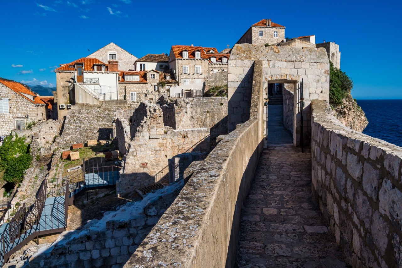 View of a walkable city wall, which is bordered on both sides by a wall. A rectangular gate within the city wall can be seen in the background, behind which are several old buildings with red-tiled roofs. On the left of the picture are some ruined remains of walls through which a fenced path leads. The sea can be seen in the back-right corner of the picture.