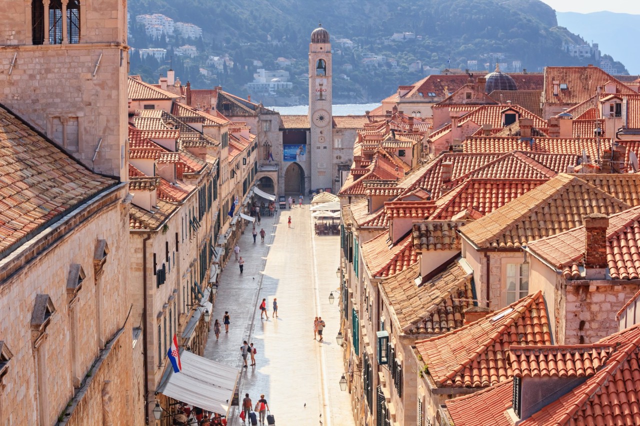 View from above into the pedestrian zone of an old town, which runs towards a narrow rectangular tower. There are many people on the street and an awning is visible on the left of the picture. The buildings on both sides of the pedestrian zone have striking, angled red-tiled roofs. On the left, a higher part of the building, presumably a tower, rises out of the picture. A rocky coastline can be seen blurred in the background.