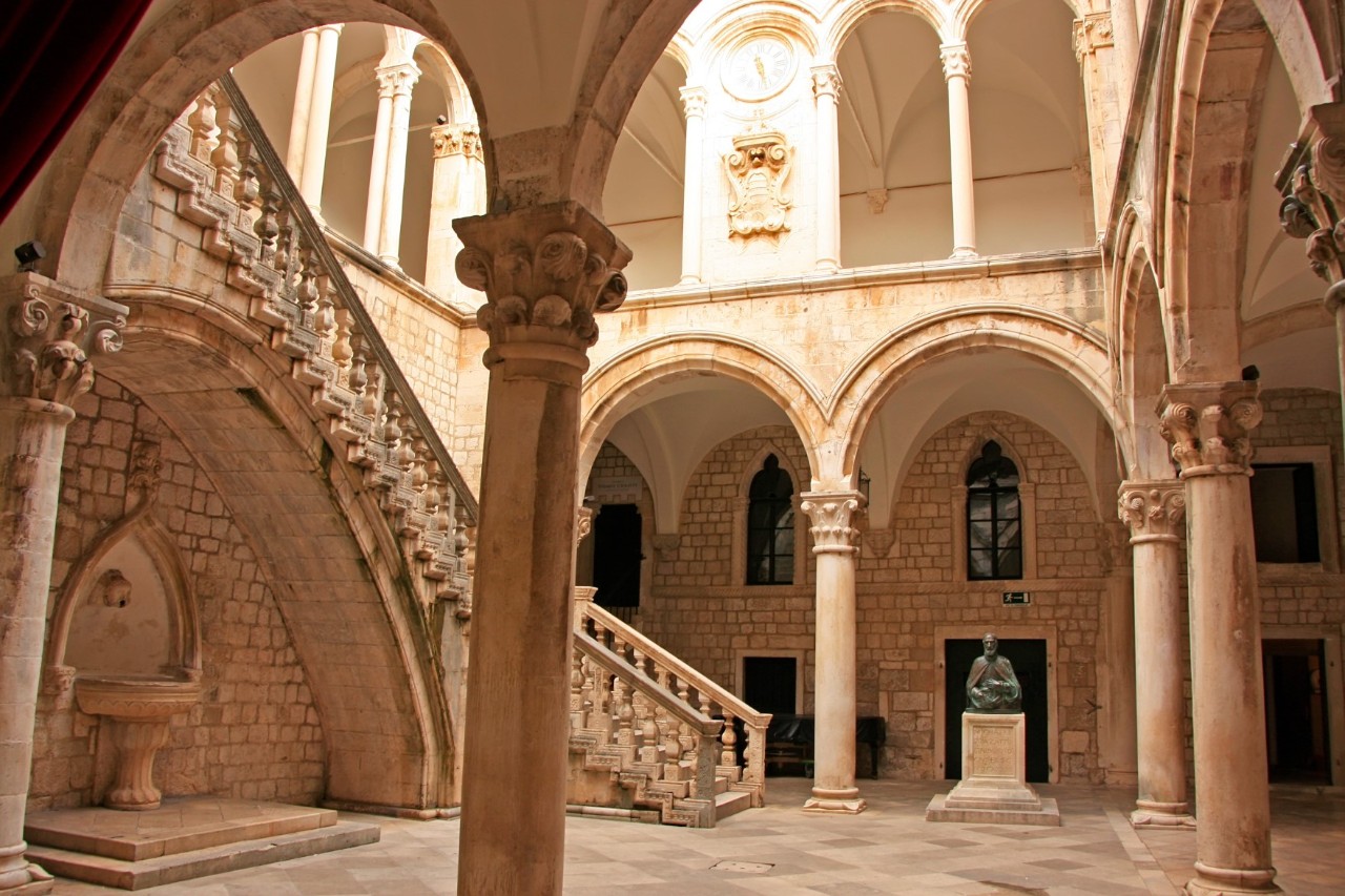 View into the inner courtyard of a light-coloured building, into which the sun is shining from above. Spread over two floors, numerous ornate round arches and columns can be seen and a curved staircase with stone balustrades leads upwards. On the left is a small water basin against the wall.