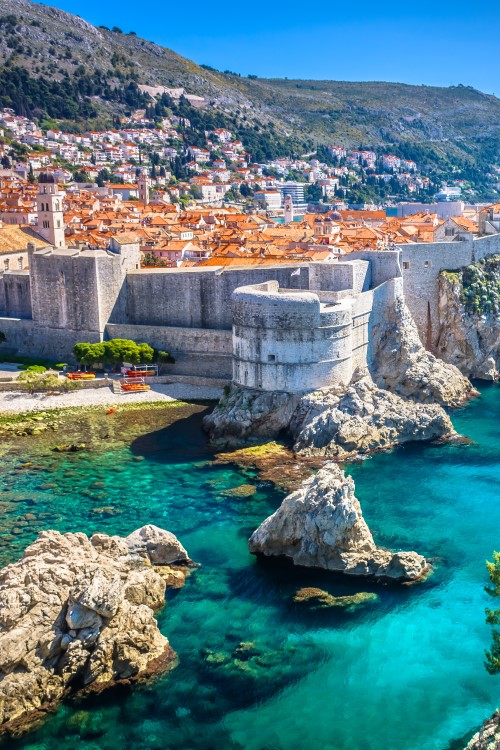 View of a city by the sea, built on a steep cliff and surrounded by a massive city wall. Numerous red roofs can be seen. There are two large fortifications within the city wall. Several small rocks rise out of the turquoise sea in front of the city. A green-covered cliff towers out of the picture on the right.