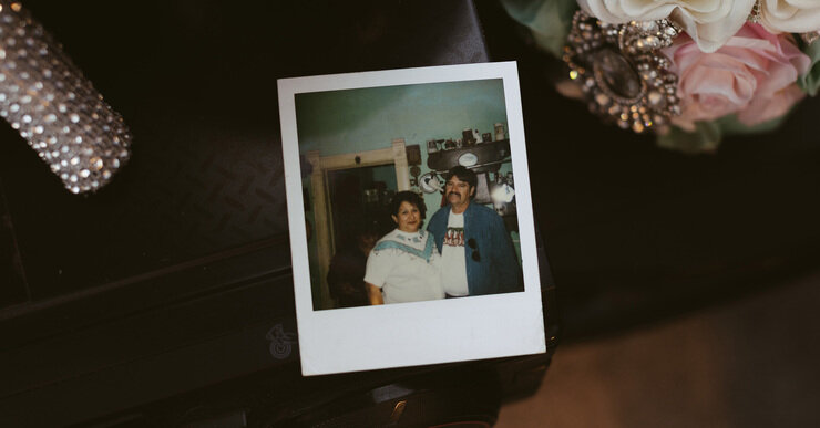 A Polaroid is on a black table. It shows Armando Navejas, a medium light-skinned man with a mustache, standing with his wife Josephine, a medium light-skinned woman. 