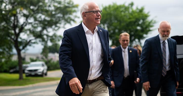 Tim Walz, a White man with white hair and glasses, wearing a navy suit jacket and khaki pants, walks outside with two men behind him to his left.  