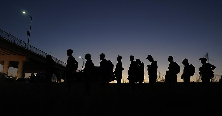 Silhouettes of about 11 people stand in a line outside. An overpass bridge and parked cars can be seen in the background.