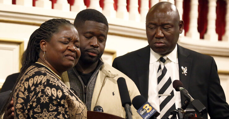 Caroline Ouko, a Black woman, and Leon Ochieng, and Ben Crump, two Black men, stand before a podium.  Ouko and Ochieng have their eyes closed and have emotional expressions on their faces.  