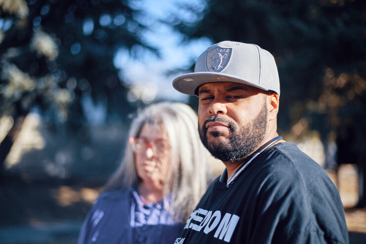 Thirty-two-year-old Anthony Kent, wearing a sports cap and black shirt, stands at a park. His mom, Teri Quintana, stands behind him on his left. 