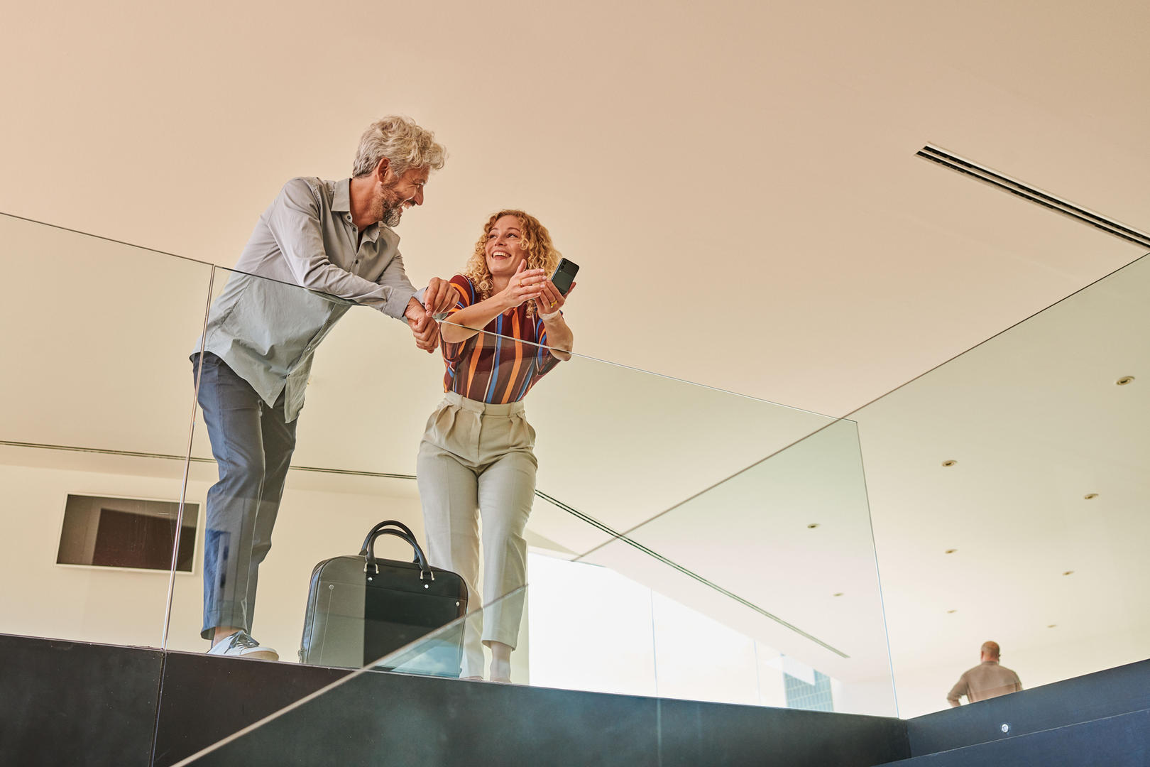 Ein Mann und eine Frau stehen oben an einem Glasgeländer und lachen. Die Frau hält ein Handy in der Hand.