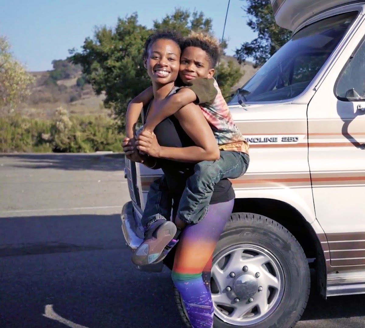 A young woman smiles at the camera while a boy ride piggyback on her. They're standing in front of the hood of a recreational vehicle parked on the side of a road.