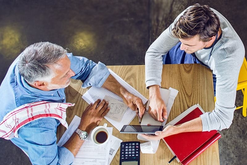 birds eye view of 2 men sitting at a table with books, and looking at an ipad