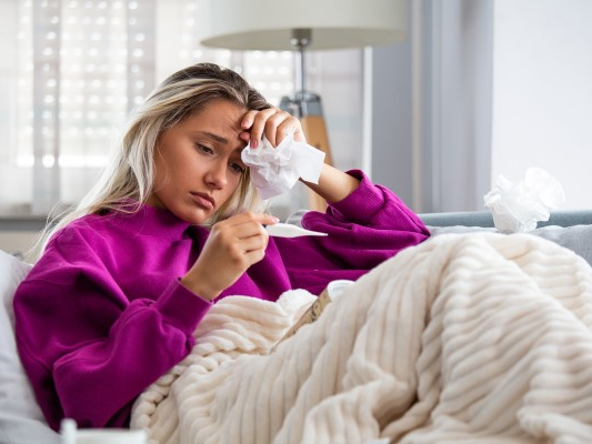 young woman suffering from COVID, sitting on the couch with tissues and a thermometer