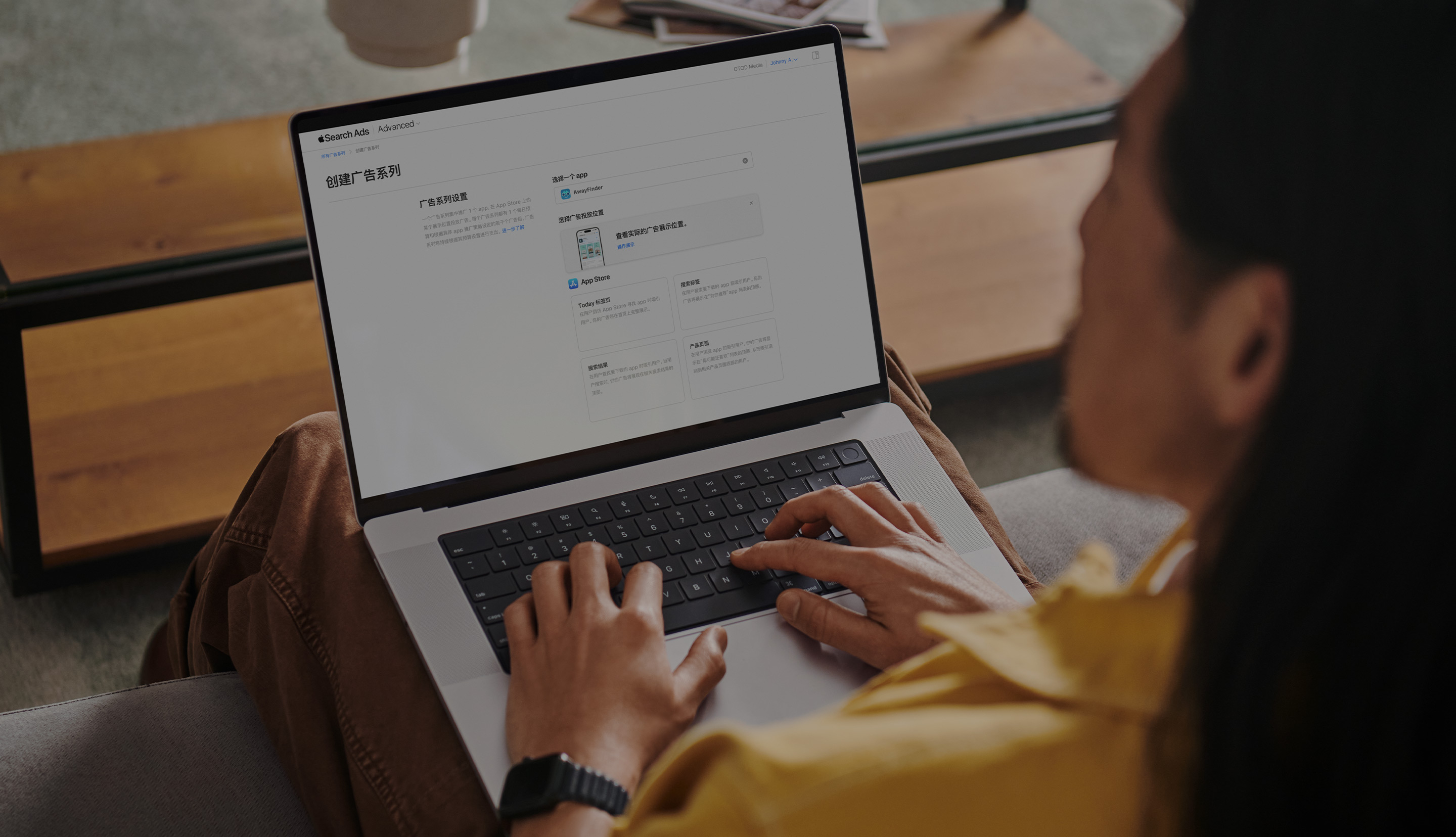 一位广告主正在使用 MacBook 工作。电脑屏幕上打开了 Apple Search Ads“创建广告系列”页面，其中显示四个可用的广告展示位置：Today 标签页、搜索标签、搜索结果和产品页面。