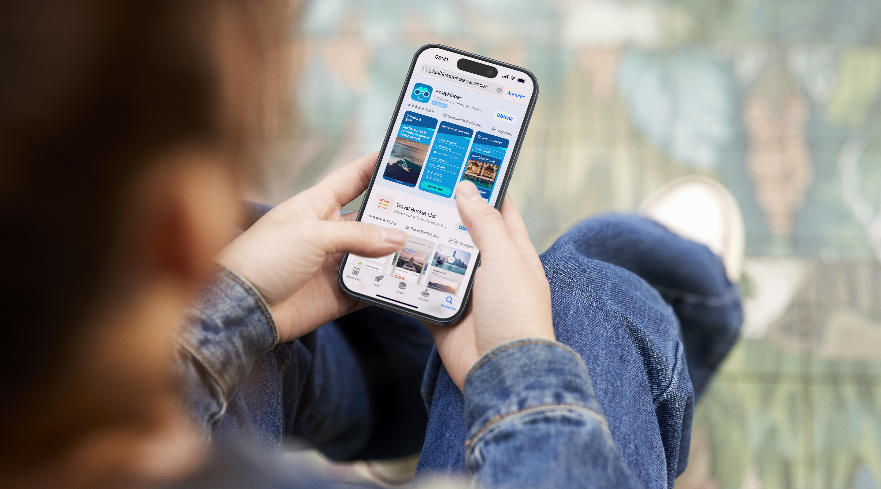 Deux mains tenant un iPhone. L’App Store est ouvert sur une annonce de l’app fictive AwayFinder dans les résultats de recherche, avec les termes « planificateur de vacances » saisis dans le champ de recherche.