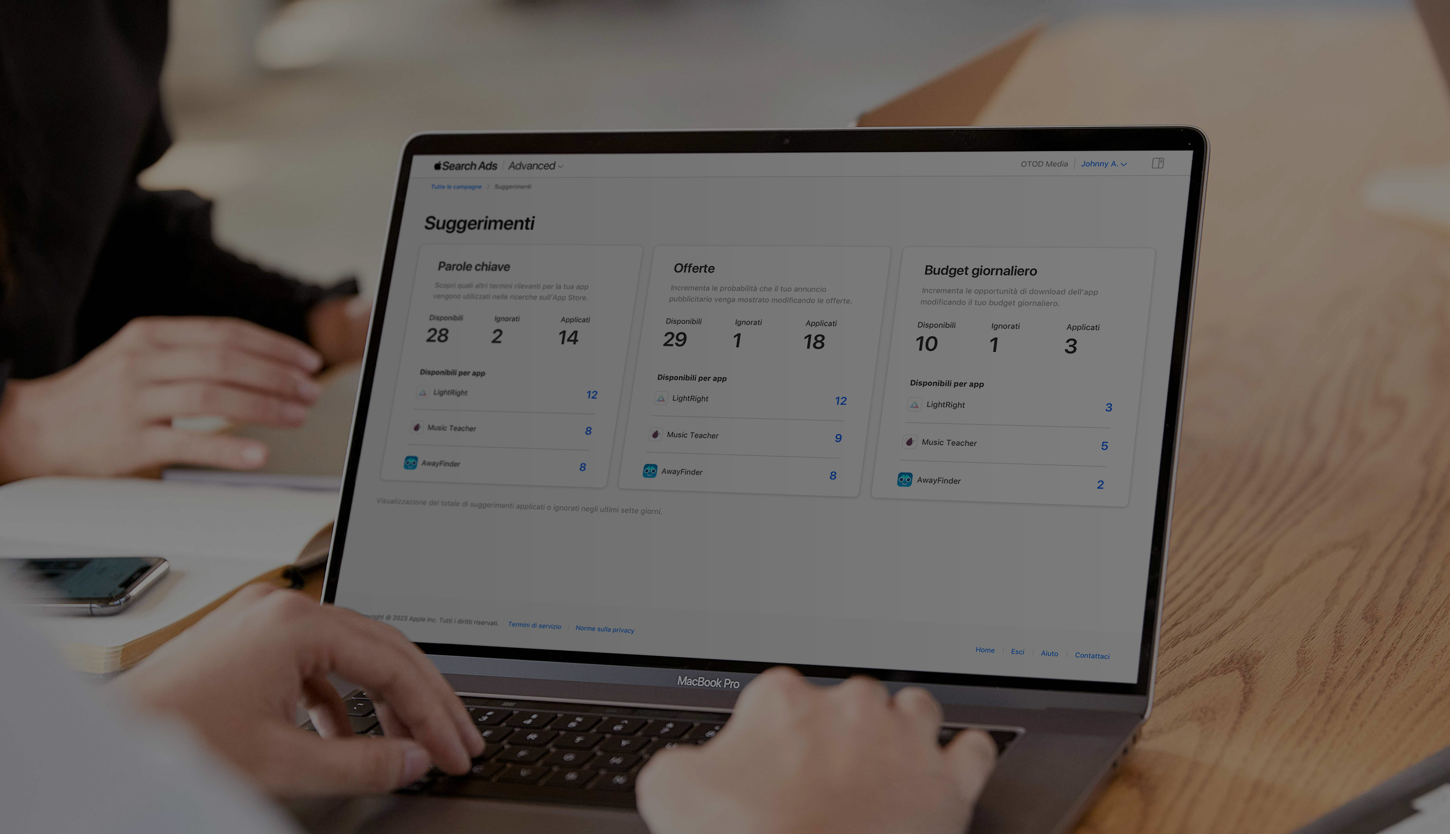 Primo piano di due mani che digitano sulla tastiera di un MacBook. La schermata è aperta sulla pagina Suggerimenti in Apple Search Ads e mostra i suggerimenti organizzati per parole chiave, offerte e budget giornaliero.